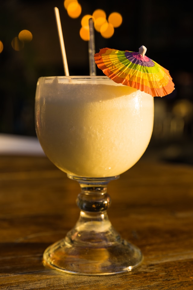A clear drinking glass containing frothy, pale yellow liquid garnished with a tiny, rainbow-colored umbrella