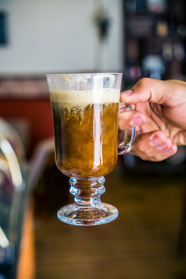 Hand holding a clear glass mug with brown, frothy drink inside