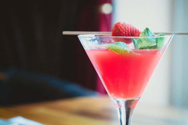 A clear glass containing pink colored liquid garnished with a piece of strawberry and mint leaves