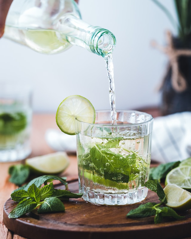A bottle pouring clear liquid into a clear glass with mint leaves inside