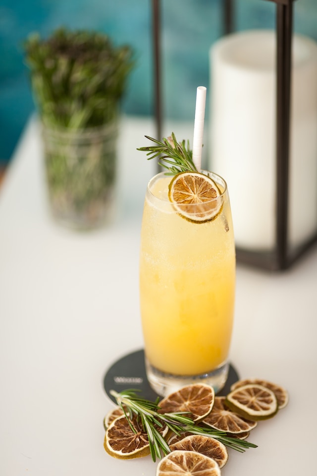 Tall clear glass with yellow liquid inside, garnished with herbs and a slice of dried lemon 
