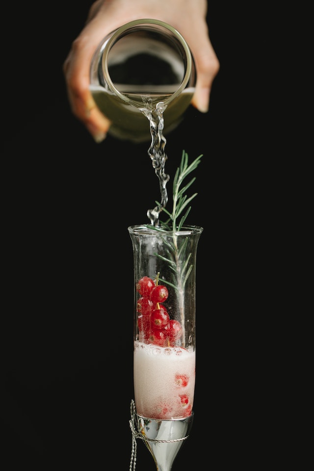 Hand pouring clear liquid into a tall clear glass containing berries and herbs