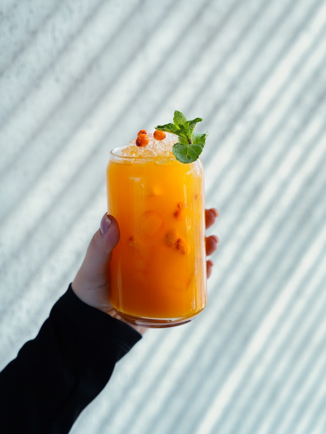 Hand holding a clear drinking glass containing eyes, fruits, and an orange liquid garnished with mint leaves