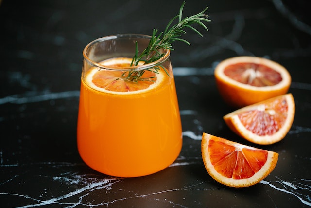 Orange liquid inside a clear glass topped with a slice of orange and rosemary