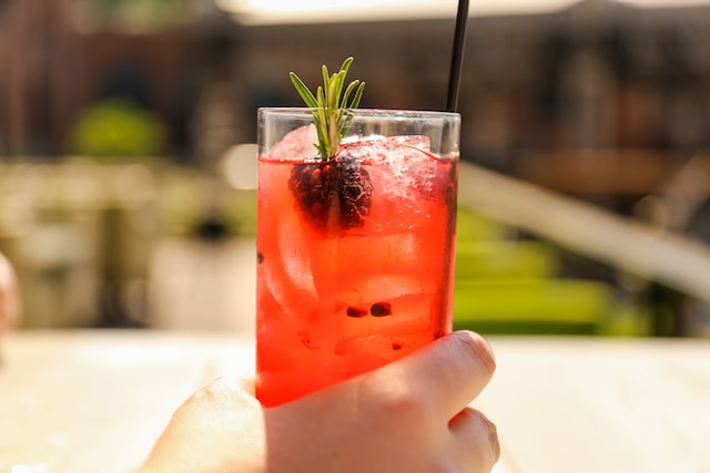 Hand holding a clear glass filled with reddish liquid ice, a sprig of rosemary and a blackberry