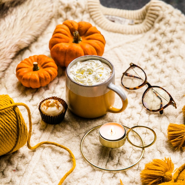 A mug of pumpkin-flavored drink with cream on top, and a cupcake on the side surrounded by a lit candle, a pair of eyeglasses and some pumpkins