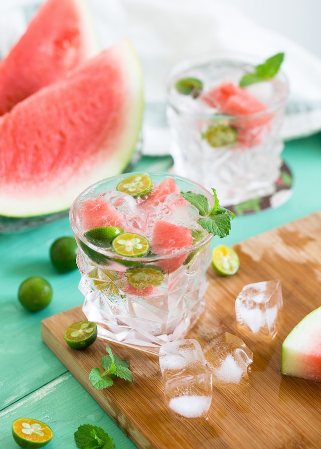 Clear beverage in a clear glass with ice cubes, watermelon cubes , basil leaves and lime